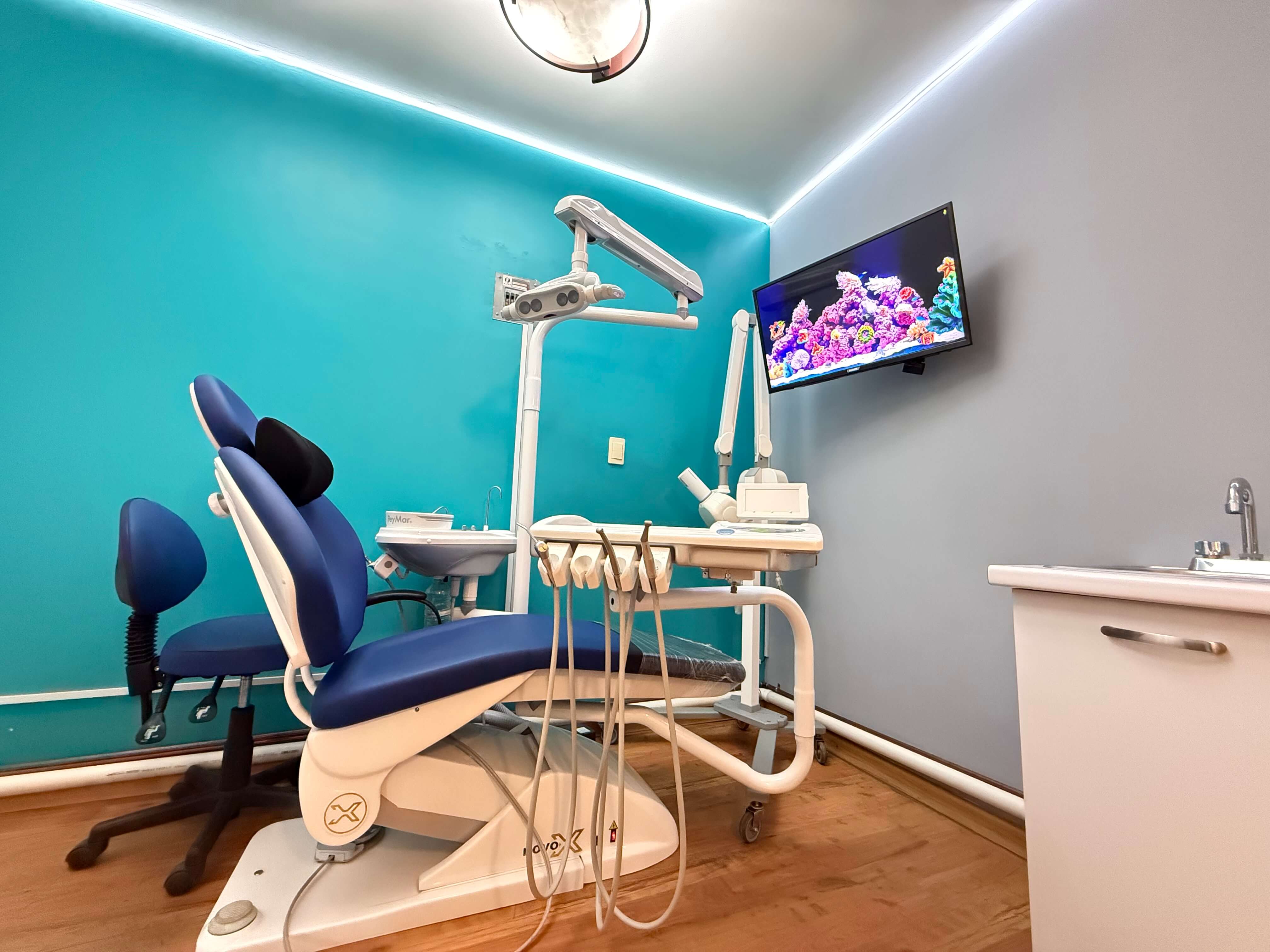 Consultorio dental con sillón odontológico azul, equipo dental, pantalla con imágenes de arrecifes y muebles blancos.